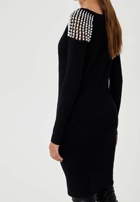 Robe en maille noire avec manches longues et détails structurés uniques aux épaules ornés de perles. Coupe ajustée avec une silhouette mi-longue.