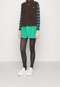 Braune Fleece-Weste mit Reißverschluss, grüner Jeansrock, gestreiftes Langarmshirt und schwarze Strumpfhosen; weiße strukturierte Slipper.