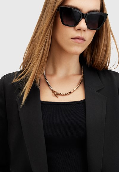 Des lunettes de soleil noires surdimensionnées, un collier en chaîne or et argent avec un fermoir, porté sur un haut débardeur noir à côtes et un blazer noir.