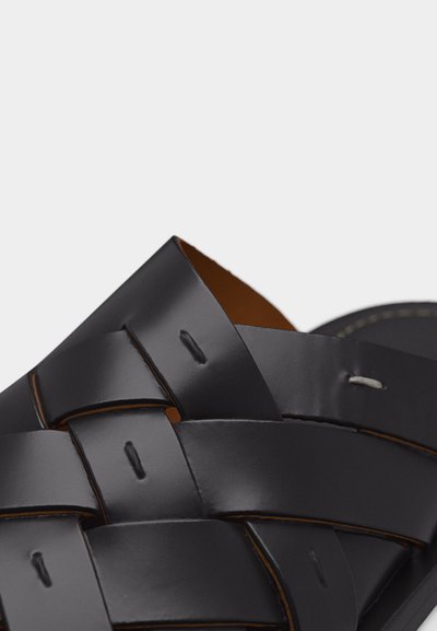 Gros plan sur une sandale en cuir noir avec des larges sangles entrelacées et un détail de couture visible sur un fond uni.