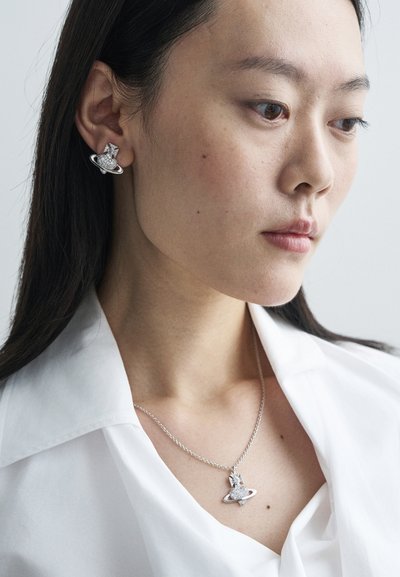 Sølvøredobber med himmelsk design og hjerte-detalj, kombinert med et sølvkjede med en lignende anheng, båret over en hvit skjorte.