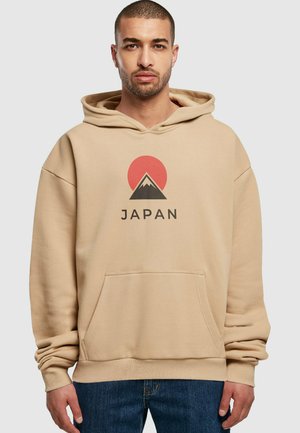 Beige mikina s kapucňou, ktorá má na prednej strane grafiku hory a slnka so slovom "JAPAN" pod ňou, vyrobená z mäkkého materiálu a s klokankovou vreckom.