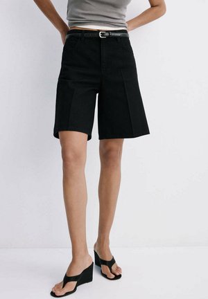 Person iført sorte shorts med høj talje og bælte, grå ærmeløs top og sorte kile-sandaler, stående mod hvid baggrund.