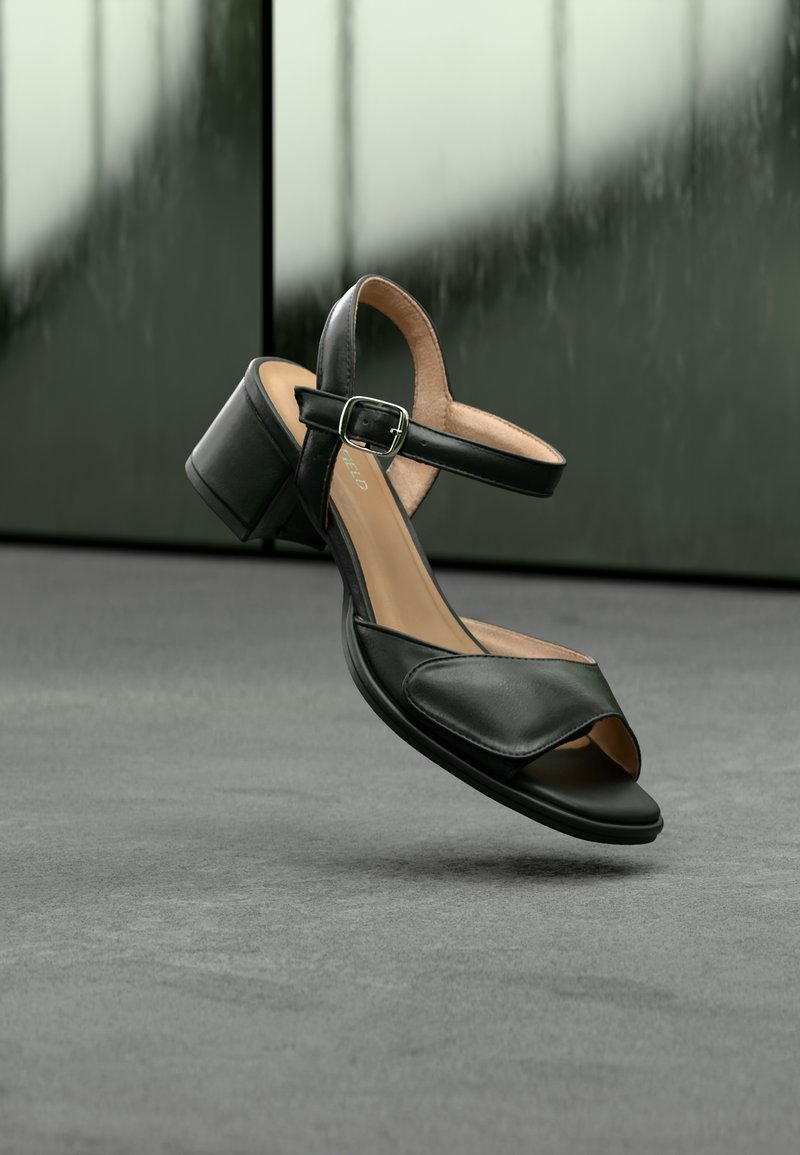 Sandale noire à bout ouvert avec bride à la cheville et petit talon bloc, présentée sur une surface grise avec un arrière-plan sombre flou.