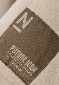 Braunes Stoffetikett mit der Aufschrift "FUTURE ICON ISSUE DESIGN NO 4002 PREMIUM", das auf beige Fleece-Material genäht ist.