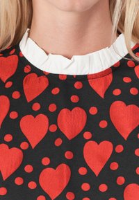 Tela negra con un patrón de corazones rojos y lunares rojos, con un cuello de volantes blanco. El acabado texturizado añade profundidad al diseño.
