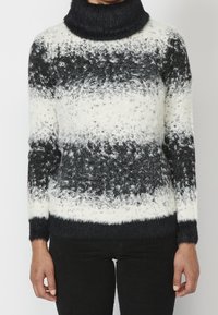 Schwarz-weißer, fuzzy gestreifter Rollkragenpullover, getragen mit schwarzen Hosen, die am Hals und mid-thigh gekürzt sind, Hände entspannt an den Seiten.