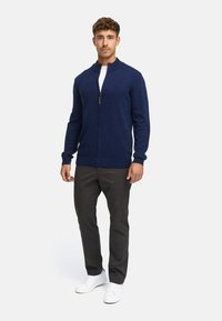 Navy zip-up cardigan in soft knit fabric, featuring a high collar, ribbed cuffs, and a brown zipper pull. Paired with dark pants and white shoes.