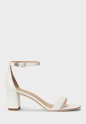 Sandale à bout ouvert en cuir blanc avec talon bloc, bride à la cheville et boucle argentée portant des initiales, sur un fond clair uni.