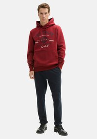 Felpa con cappuccio rosso con testo grafico, tasca a marsupio, polsini e orlo a coste abbinata a pantaloni navy e scarpe nere stringate.