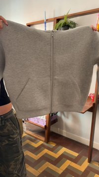 Felpa grigia con cerniera frontale e tasche laterali, realizzata in tessuto morbido, con design semplice e spalle larghe e arrotondate.