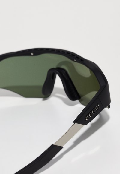 Óculos de sol pretos com lentes verdes, com um design desportivo e uma armação texturizada. Detalhes em prateado e um logotipo na haste.
