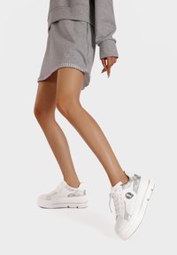 Sneakers bianchi con un accento testurizzato argento e logo. Suola spessa, superficie liscia, punta arrotondata e design con lacci. Cuciture a contrasto sui lati.