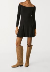 Mustikkapohja, hihaton pitkähihaisia mekko, jossa on levenevä hameosa, yhdistettynä vaaleanruskeisiin mokkasaappaisiin, jotka ulottuvat polveen ja joissa on koristeellista tikkausta.