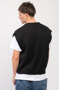 Svart strikket uten ermer genser med ribbestrikkede kanter, brukt over en hvit t-skjorte. Synlig tekstur og løst tilpasset design.