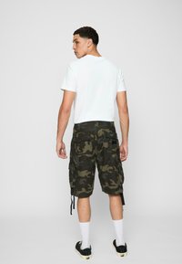 Tricou alb din bumbac, pantaloni scurți cargo verzi cu camuflaj și buzunare, adidași negri, șosete albe. Văzut din spate, fundal minimalistic.