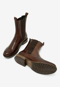 Bottines en cuir marron avec panneaux élastiqués sur les côtés, texture lisse, tirettes, et semelle en caoutchouc crantée. Construction robuste avec un design classique.