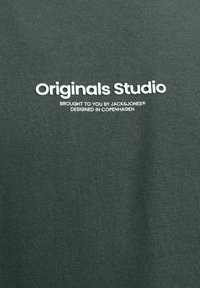 Tessuto verde scuro con testo bianco "Originals Studio" e testo più piccolo "Portato a te da Jack & Jones, progettato a Copenaghen."