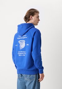 Sudadera azul de mezcla de algodón, con un bolsillo frontal y capucha con cordón. En la parte trasera muestra texto blanco y detalles gráficos.