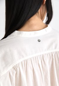 White cotton blouse featuring a gathered back design, smooth texture, and a small metallic accent at the neck.