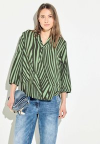 Blusa patterned verde e nera con colletto e maniche a tre quarti, abbinata a pantaloni in denim azzurro chiaro. Tessuto morbido con texture delicata.