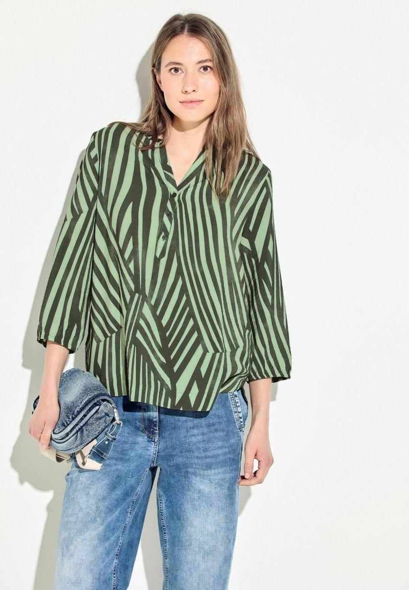 Blusa patterned verde e nera con colletto e maniche a tre quarti, abbinata a pantaloni in denim azzurro chiaro. Tessuto morbido con texture delicata.