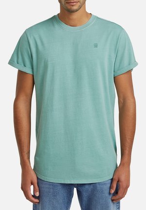 Man draagt een licht turquoise T-shirt met korte mouwen met opgerolde mouwen en blauwe spijkerbroek, staand tegen een effen achtergrond.
