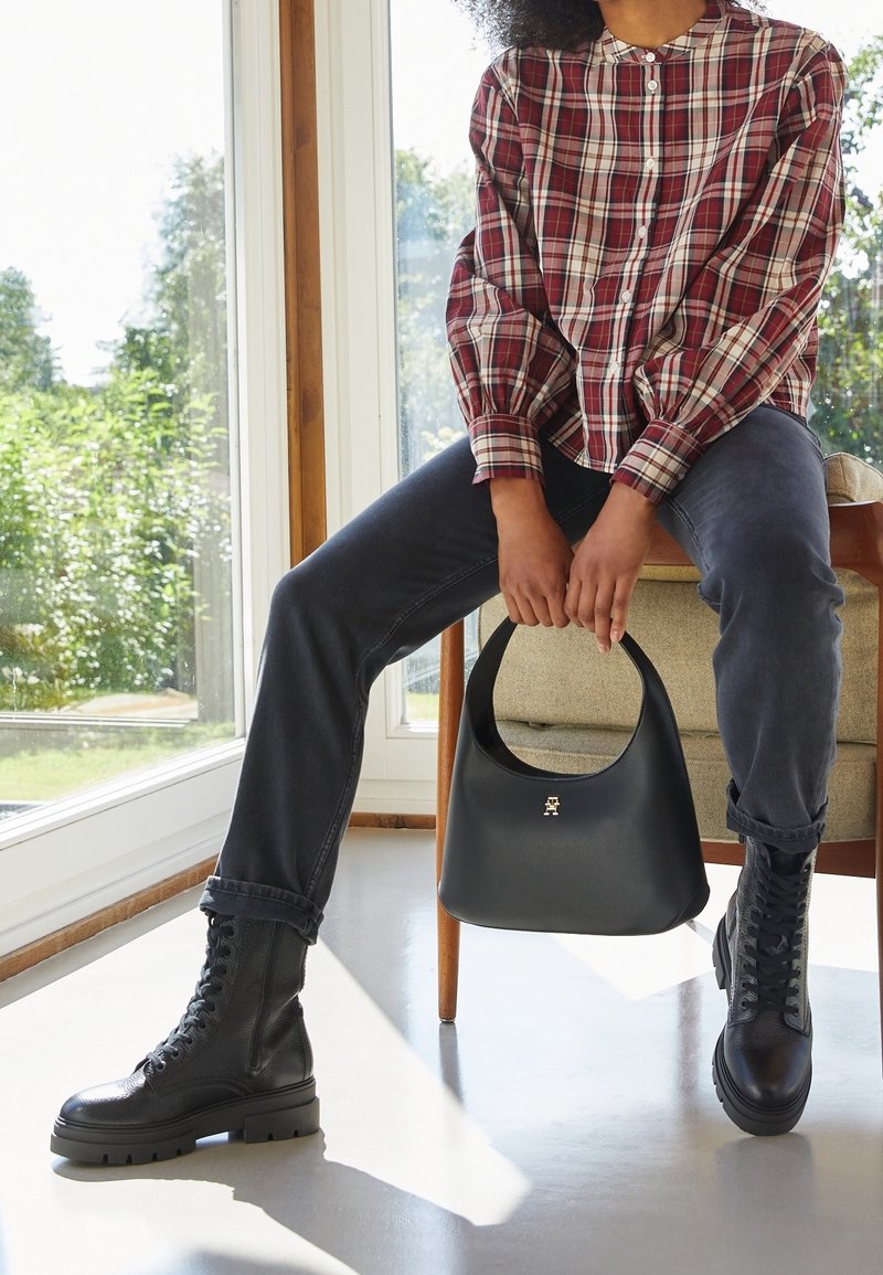 Personne portant des bottes noires à lacets et un jean foncé, assise à l'intérieur près d'une fenêtre, tenant un sac à main noir avec un fermoir doré.
