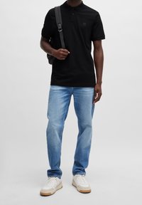 Černé polo tričko s jemným logem, kombinované se světlými modrými džíny a bílými teniskami. Tričko má design s límečkem a krátkými rukávy.