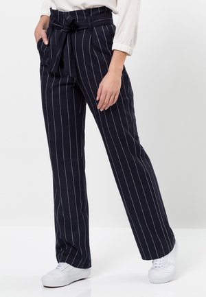 Kobieta ubrana w granatowe spodnie w prążki z wysokim stanem i wiązanym paskiem, białą bluzkę oraz białe trampki, stojąca z rękami przy kieszeniach.