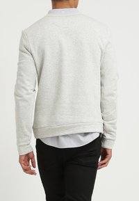 Sudadera gris con puños y dobladillo de canalé, presenta un cuello redondo y una camisa con cuello visible debajo. Diseño minimalista.