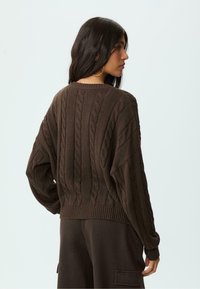 Brauner, kabelstrickartiger Pullover mit lockerer Passform, geripptem Hals- und Armabschluss, ausgestattet mit einem strukturierten Muster. Trägt dazu passende dunkle Hosen.