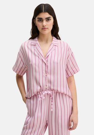 Junge Frau trägt ein passendes hellrosa kurzärmeliges Hemd mit Knöpfen und Hose mit dünnen vertikalen roten Streifen, steht mit einer Hand in der Tasche.