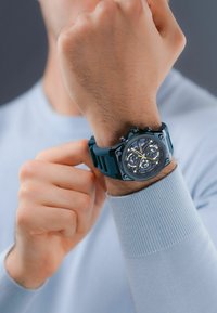 Blå gummirem klocka med en rund svart och blå urtavla, som har flera underskivor, silverdetaljer och en texturerad yta.