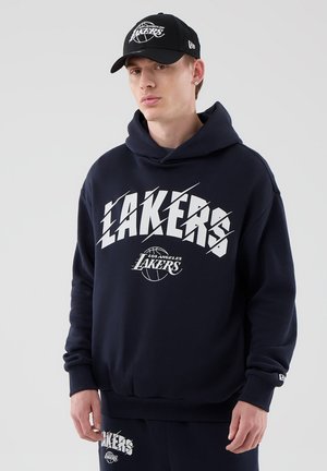 Młody mężczyzna w czarnej bluzie z kapturem Los Angeles Lakers, pasujących spodniach dresowych i czapce Lakers, stoi na jednolitym tle.