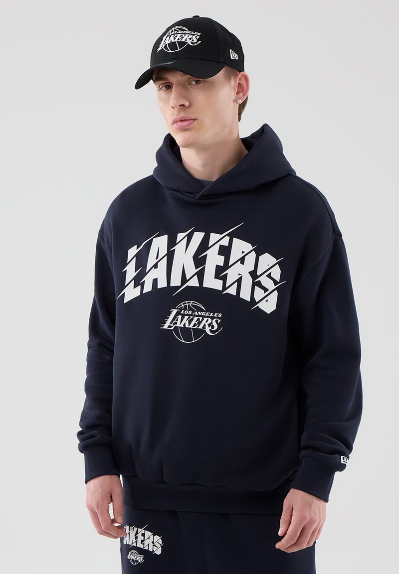Νέος άνδρας που φοράει μαύρο φούτερ Los Angeles Lakers, ταιριαστό παντελόνι φόρμας και μαύρο καπελάκι Lakers, στέκεται μπροστά σε απλό φόντο.