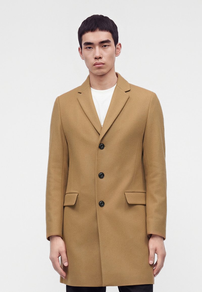 Νέος άνδρας φοράει μια ραμμένη στα μέτρα του καμηλό παλτό με τρία μαύρα κουμπιά και τσέπες με καπάκια, στέκεται μπροστά σε ένα απλό φωτεινό φόντο.