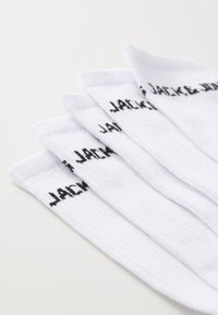 Cinq paires de chaussettes en coton blanc avec des bords côtelés, arborant le texte noir "JACK & JONES" sur le côté. Texture douce et design athlétique.