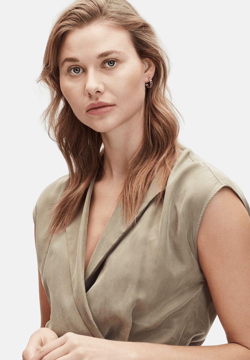 Beige, drapierter Wickeloberteil mit tiefem V-Ausschnitt, weichem Material und kurzen Ärmeln. Das Model hat welliges Haar und trägt minimalen Schmuck.