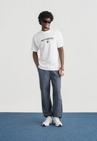 Λευκό T-shirt New Balance με μαύρο λογότυπο, γκρι παντελόνια και λευκά αθλητικά παπούτσια. Το μοντέλο φοράει μαύρα γυαλιά ηλίου σε ουδέτερο φόντο.