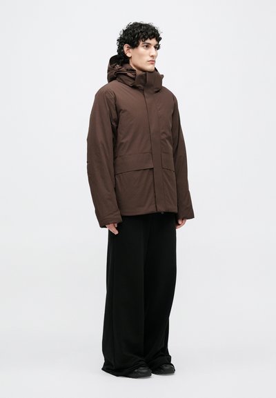 Brūna izolēta jaka ar augstu apkakli un kapuci, priekšpusē rāvējslēdzējs un divas lielas priekšējās kabatas, kopā ar vaļīgām melnām biksēm.