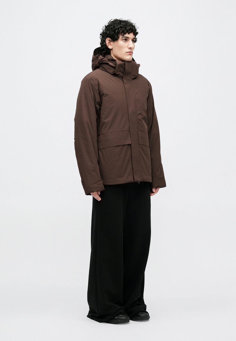 Bruine geïsoleerde jas met een hoge kraag en capuchon, voorzien van een ritssluiting aan de voorkant en twee grote voorzakken, gecombineerd met losse zwarte broek.