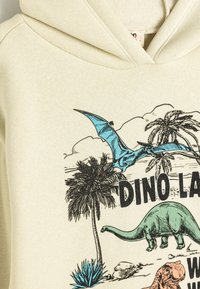 Sudadera beige con gráficos de un pterosaurio azul vibrante, un dinosaurio verde y una palmera. Textura suave y diseño de capucha con cordón.