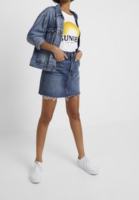 Jeansová bunda přes bílý tričko s duhovou grafikou, kombinovaná s modrou džínovou sukní s roztrhaným lemem a bílými teniskami.