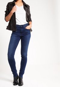 Veste en cuir noire sur un t-shirt gris, associée à un jean skinny bleu foncé et des bottines noires. Textures lisses et design ajusté.