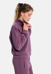 Fioletowy sweter z wysokim kołnierzem i długimi rękawami, o luźnym kroju i miękkiej fakturze. Z dodatkiem ukrytego detalu z guzikiem.