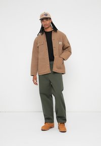 Brun foret jakke med forlommer, sort skjorte, olivengrønne bukser og brune støvler. Beige kasket med logo detaljer.