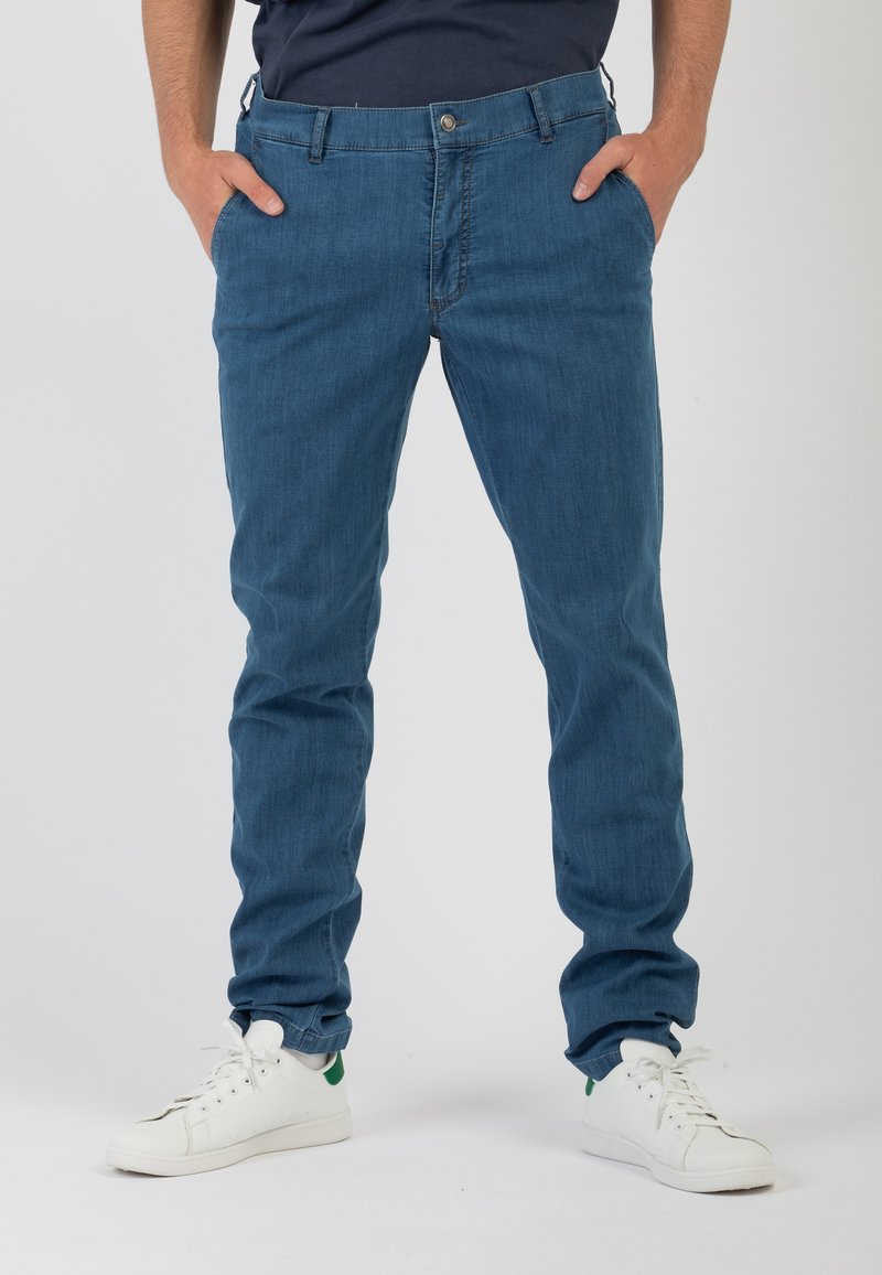 Pantaloni slim-fit in denim blu con una texture liscia, cinque tasche e zip. Abbinati a sneakers bianche con accenti verdi.