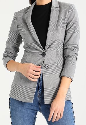 Frau trägt einen grau karierten Blazer mit zwei Knöpfen über einem schwarzen Top und blauen Jeans mit seitlichen Knopfdetails, Hände passen den Blazer an.