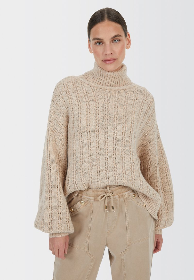 Beige geripptes Sweatshirt mit hohem Rollkragen und weiten Glockenärmeln, aus strukturiertem Strick und weichem Material. Kombiniert mit hellbeigen Hosen.